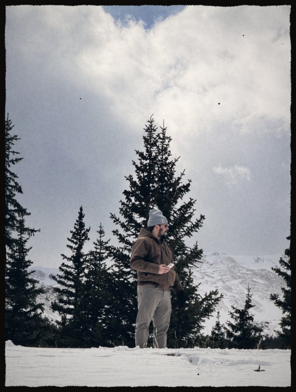 A man in a beanie standing in snow below evergreens and clouds