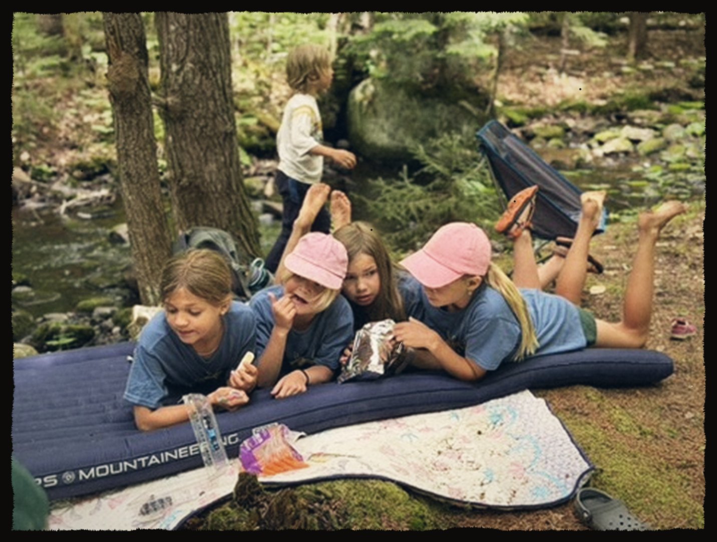 Kids sharing snacks on a sleeping pad by a creek