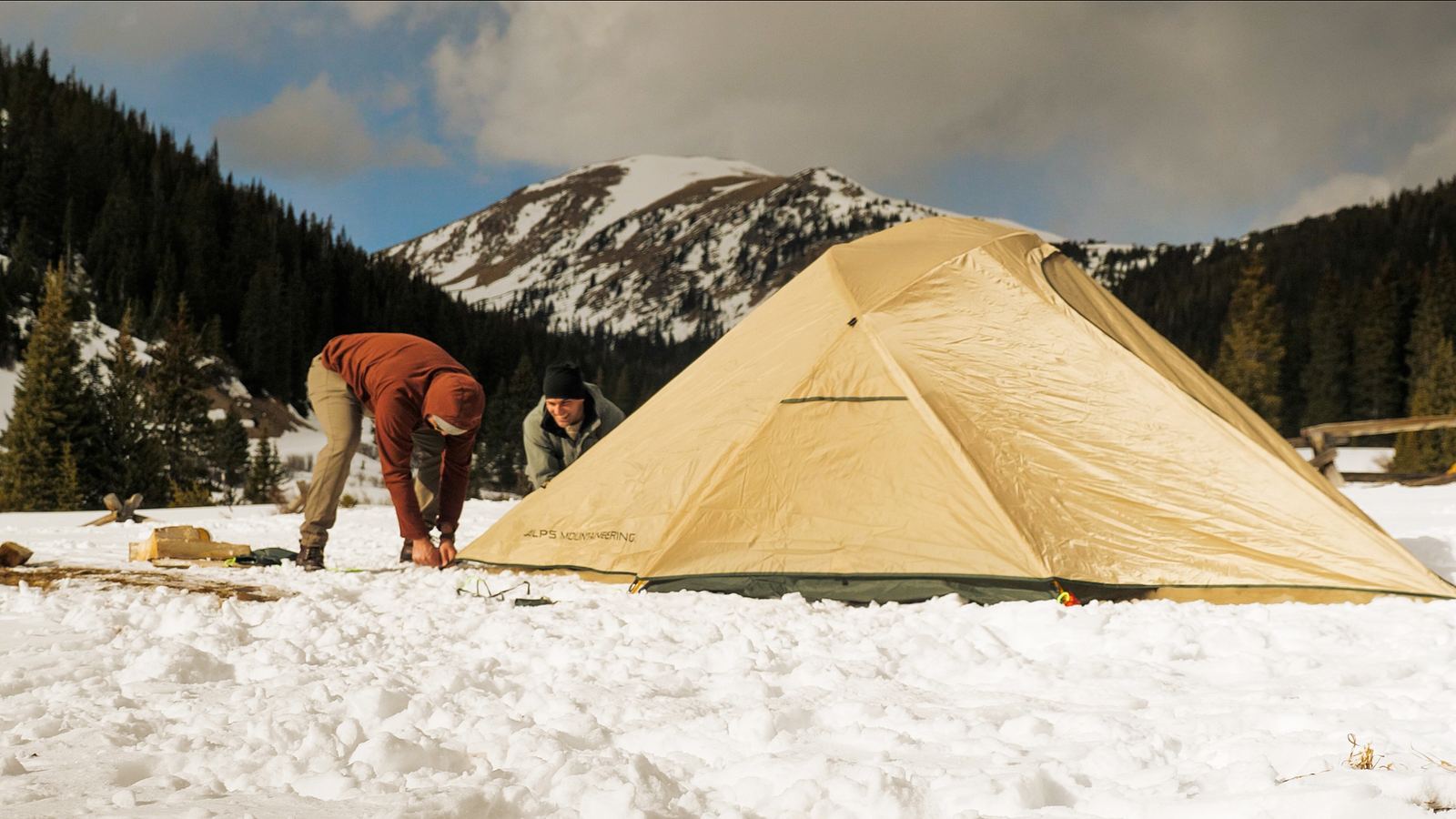 Two campers pitching a tan tent in snow below a mountain ridge